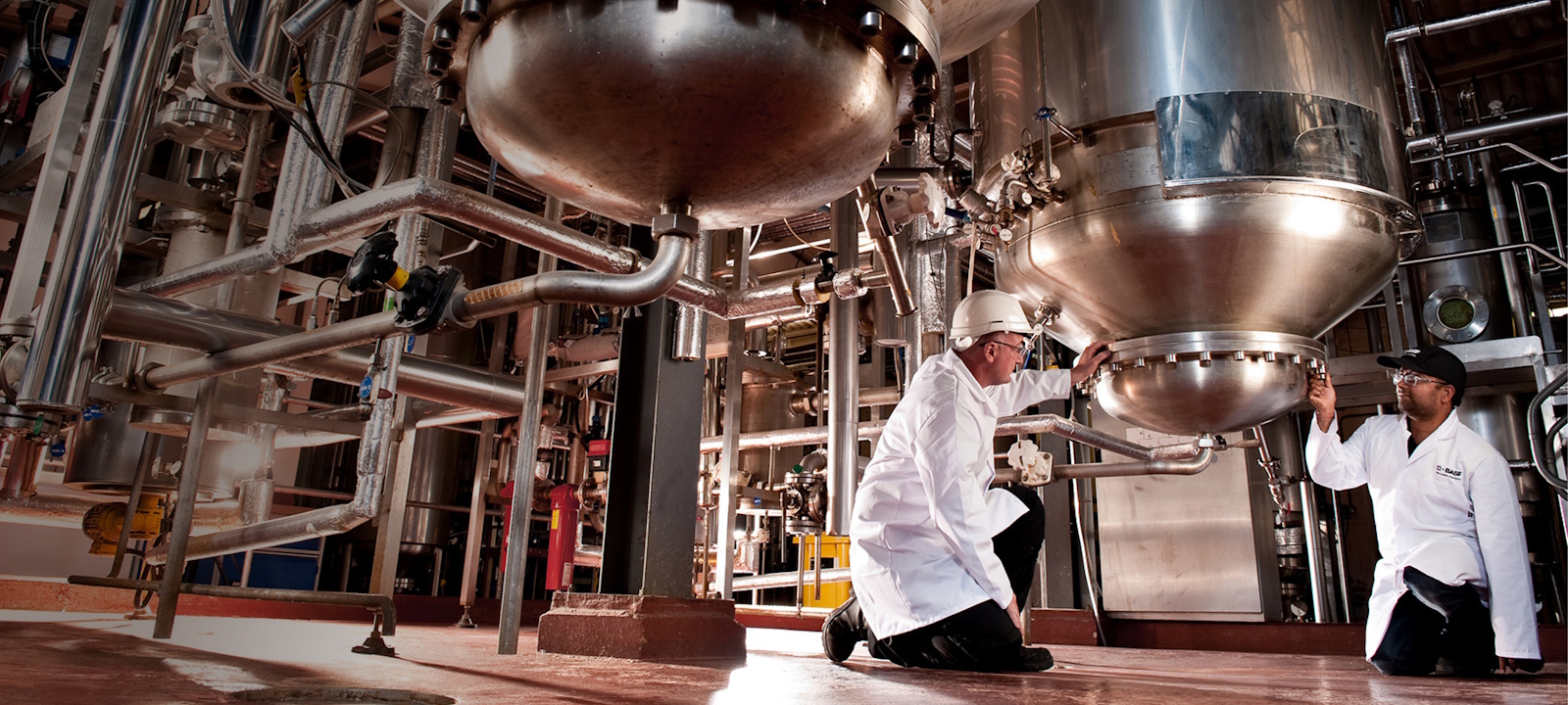 two people in white coats kneeling in front of large industrial machinery
