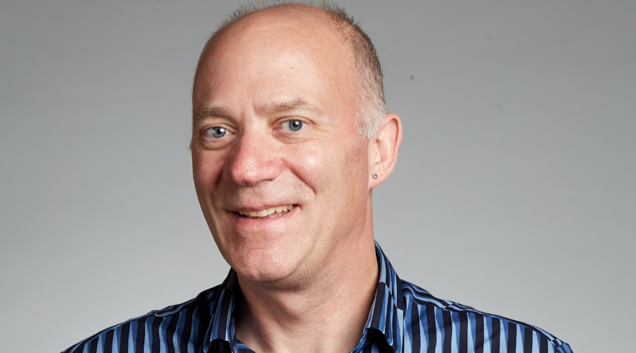 Professor John Hutchinson, wearing a navy patterned shirt and standing in front of a grey background, smiles at the camera.
