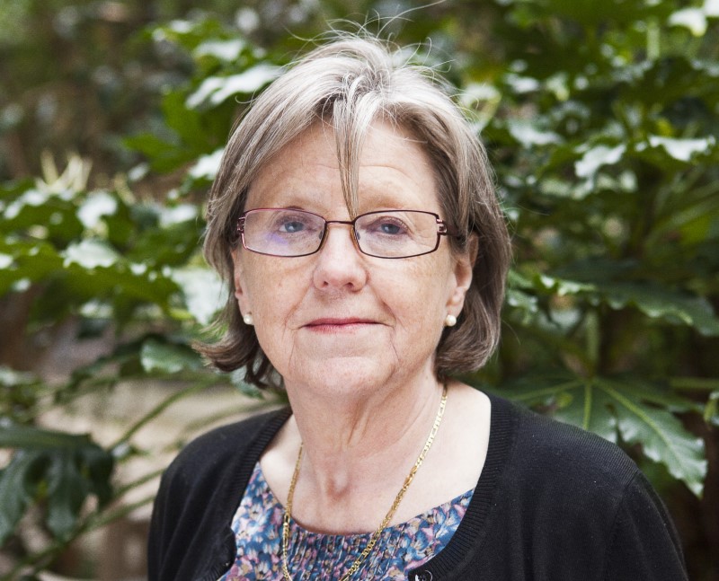 Photographic portrait of Georgina Mace FRS, by photographer Jussi Puikkonen. She is standing outside with foliage in the background. She's wearing glasses and looking in to the camera. She is wearing a necklace and a black cardigan.